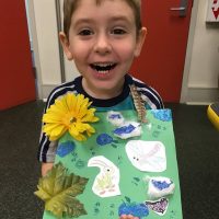 young boy smiles at camera and holds up art project