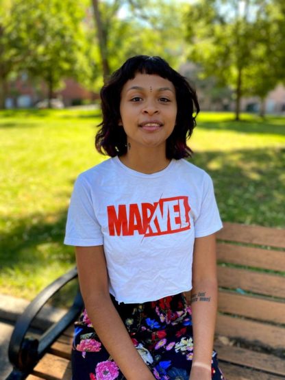 Adriana sits on a park bench in a grassy field, wearing a Marvel T-shirt