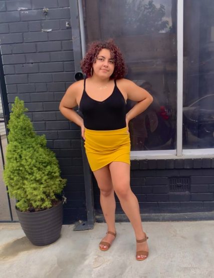 Yanni stands in front of a window, hands on hips in a yellow skirt & black top