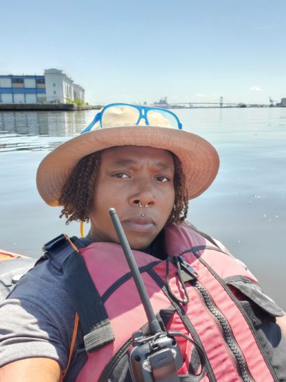 Brooklyn sits in a kayak on the Schuylkill Rive, blue glasses perched on a hat on head, a walkie talkie partially blocking their face