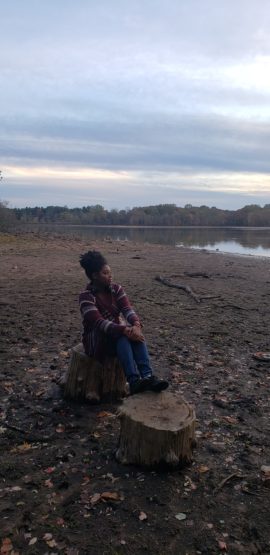 MyKyah sits on a rock on the shoreline of a body of water, gazing away from the camera