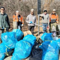 a group of volunteers pose around bags of trash they've collected in a wooded area along a trail.